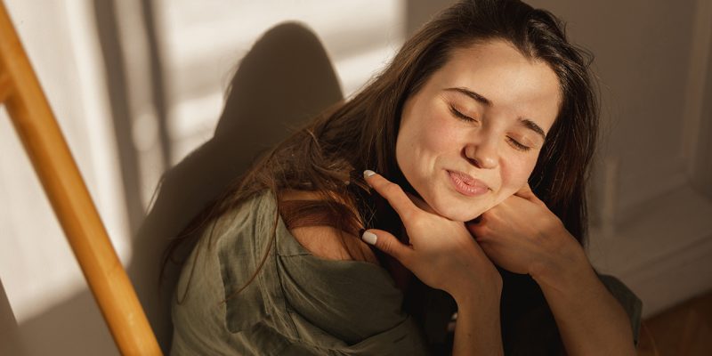 attractive-caucasian-young-girl-smiles-modestly-camera-closing-her-eyes-from-sun-brunette-sits-light-room-against-white-wall-leisure-lifestyle-beauty-concept