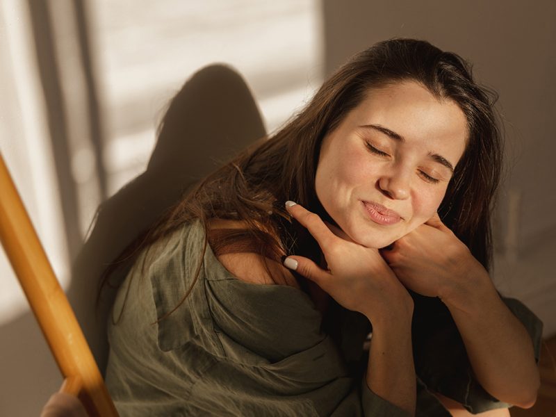 attractive-caucasian-young-girl-smiles-modestly-camera-closing-her-eyes-from-sun-brunette-sits-light-room-against-white-wall-leisure-lifestyle-beauty-concept